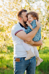 Dad hugs and kisses his little daughter on a walk in a blooming spring garden.