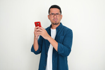 Adult Asian man looking at the camera with disappointed expression while using his phone