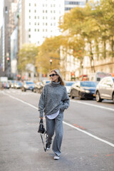 full length of blonde woman in sunglasses and trendy outfit holding handbag while walking on street of New York city