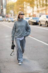 full length of blonde woman in sunglasses and grey outfit holding handbag while walking on street of New York city