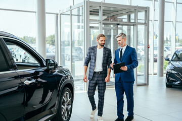 Male dealer with a customer showing new crossover holding tablet with documents standing in the showroom.