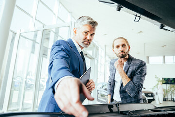 Handsome salesperson with client in the car showroom near open hood.