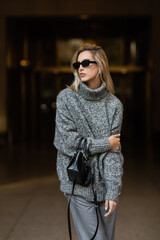 stylish woman in sunglasses and grey sweater standing with black handbag in New York