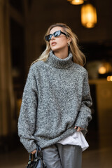 blonde woman in stylish sunglasses and grey sweater standing with black handbag in New York
