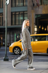 full length of stylish young woman with blonde hair walking near yellow cab in New York