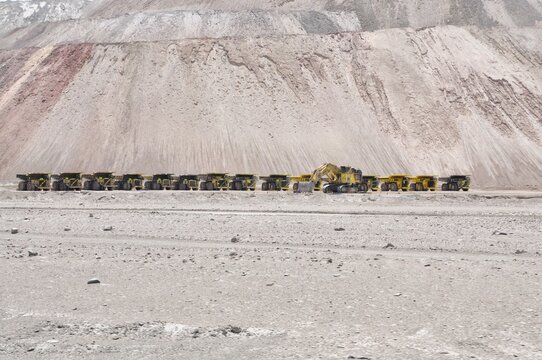 Huge mining trucks are parked in a row in front of a big hill. Mining trucks in Atacama desert, Chile. Lithium mining industry in Chile. Big mining trucks and diggers look like toys in the distance 