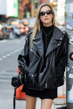 stylish young woman in leather jacket and sunglasses standing with hands in pockets in New York