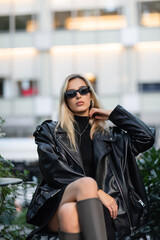 Obraz na płótnie Canvas young blonde woman in black leather jacket and trendy sunglasses sitting in outdoor cafe