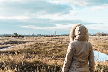 Fototapeta premium Back view of woman wearing winter clothes in front of grassland wilderness and factories on the horizon - Winter landscape with industries on background - Activism and environmental protection concept