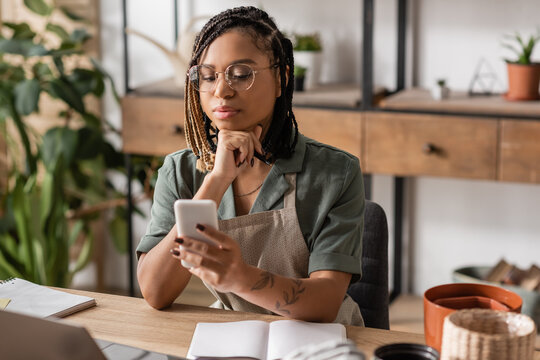thoughtful african american florist in eyeglasses looking at cellphone near blank notebook in flower shop