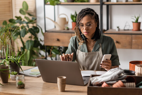 focused african american florist sitting at workplace with smartphone and notebook in flower shop - Powered by Adobe