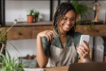 cheerful african american florist with trendy hairstyle and eyeglasses looking at mobile phone in flower shop