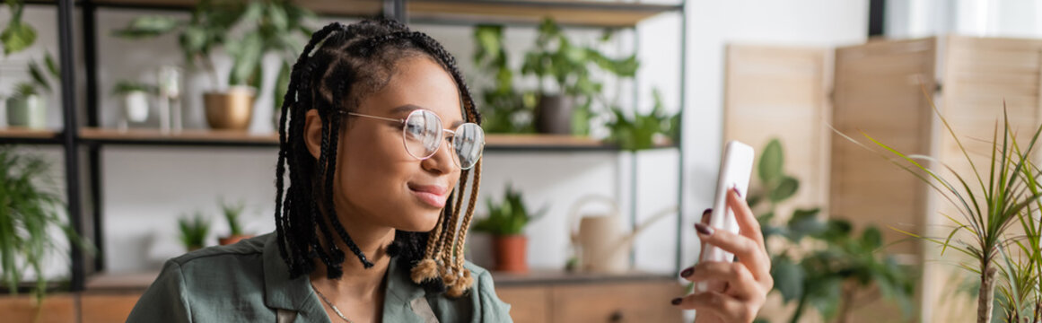 stylish african american florist with dreadlocks and eyeglasses looking at smartphone in flower shop, banner