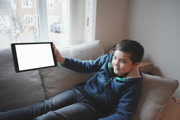 Happy Child with tablet computer. Portrait Kid Holding And Showing Digital Tablet With Empty Screen  Mockup, Young boy sitting on sofa relaxing and playing with touch pad at home © Anchalee