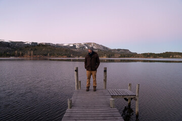 The placid lake at nightfall. Magical view of a man standing in the wooden dock with the mountains...