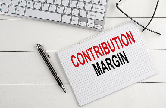 CONTRIBUTION MARGIN text on notebook with keyboard , pen glasses on white wooden background