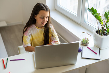 Photo of positive kid girl study remote sit desk table use laptop have online communication lesson with tutor writing report in house indoors.