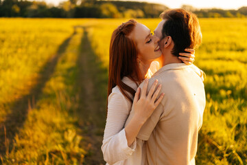 Fototapeta premium Smiling redhead young woman with closed eyes and unrecognizable man in love hugging, kissing, stroking standing together on beautiful green meadow, in summer evening during golden sunset.