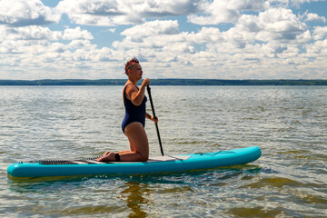 A woman in a swimsuit with a short haircut on a SUP board with a paddle in the lake against the background of white clouds.