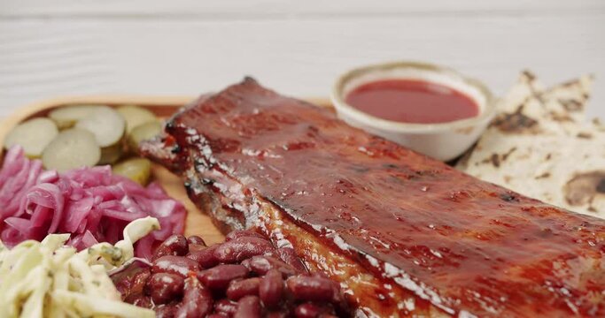 A large Plate that includes Baby Back Ribs with cherry chipotle sauce. cooked slowly on smoker, sauce, BBQ beans, coleslaw, pickled onions, pickled cucumbers, Rotating on isolated white background