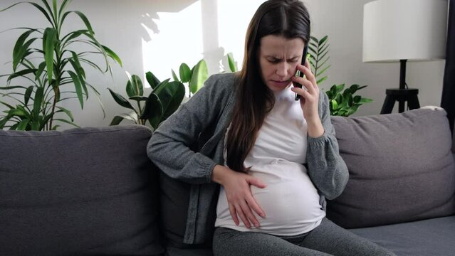 Close up of unhappy caucasian young pregnant woman feeling pain sitting on sofa in living room, touching belly and having call. Pregnancy, motherhood, expecting baby and spending time at home
