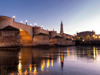 View of the tower of La Seo de Zaragoza cathedral with the medieval stone bridge over the Ebro river in the foreground during sunset in winter.