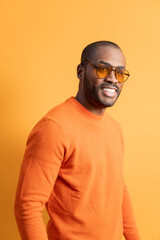 Fototapeta premium A dark-skinned adult male gazes out 45 degrees while posing against an orange wall wearing matching sunglasses. African american fashion concept.