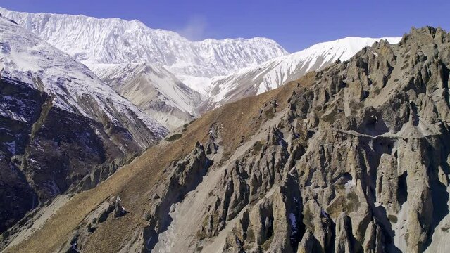 Flying along steep rugged cliffs in Manang Nepal with snow capped mountains in the distance and trail below.