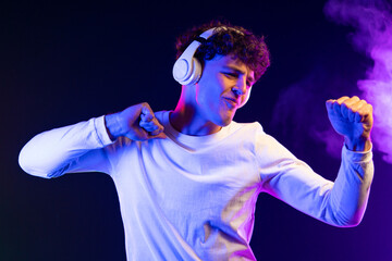 Positive man listening music with headphones, dancing on dark neon background. Stylish student guy...