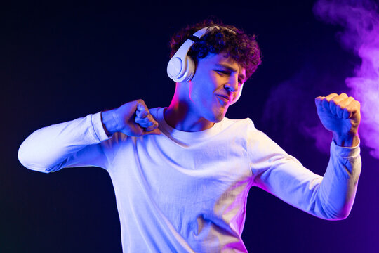 Positive man listening music with headphones, dancing on dark neon background. Stylish student guy enjoying life, active energy, inspired dance concept
