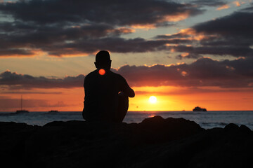 Man sitting alone on beach rocks looking at the sunset, lost in thought