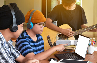 Group of young asian teenagers sitting together inside the room working and practising school project about the music subject through laptop and playing quitar, too, soft and selective focus.