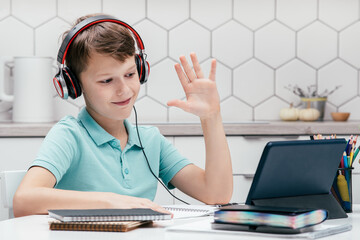 Portrait of young preteen wonderful boy wearing headphones, sitting at desk near notebooks, looking at screen of tablet, having online lesson, raising hand, greeting. Remote education, e-learning.