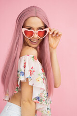 Girl in pink heart-shaped glasses with pink hair looks at the camera with her glasses slightly lowered and smiling Pin up smile Emotions Pink background Vertical frame