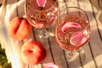 Glasses of delicious rose wine with petals and peaches on white picnic blanket, above view