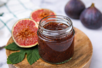 Glass jar of tasty sweet fig jam and fruits on white table, closeup