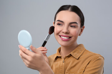 Happy woman with cosmetic pocket mirror applying makeup on light grey background