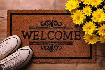 Doormat with word Welcome, stylish sneakers and beautiful flowers on wooden floor, flat lay