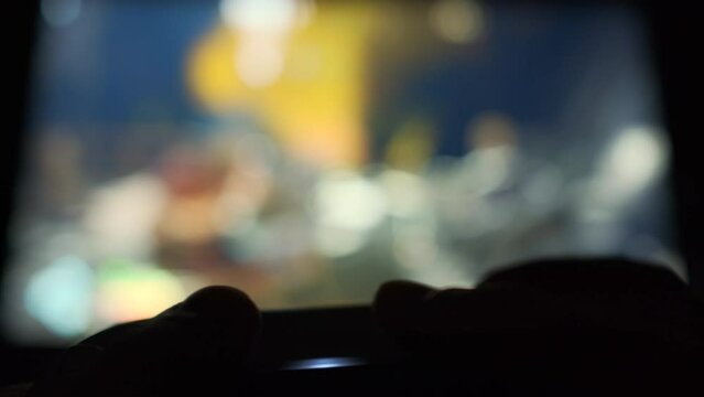 Close up view of persons hand holding a video game controller with bright blue lights playing an intense and colorful video game on big screen TV in dark living room