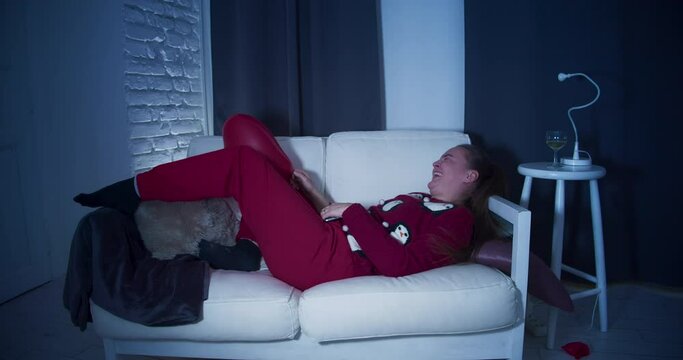 A young woman lying in sweatpants on a couch with a mobile phone,  flirts online with her boyfriend on Valentine's night.