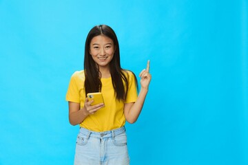 Happy Asian woman holding a phone with a yellow case on a blue background in a yellow T-shirt with a smile and teeth pointing upwards