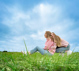 Two young girls are resting, relaxing, sitting on the grass by the sea. Hugging and smiling. Comfortable, casual clothes. Summer day.