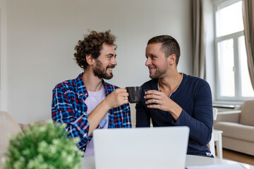 Lovely gay couple laughing together while sitting in their living room at home. Two romantic young male lovers having fun surfing the internet indoors. Young gay couple living together.