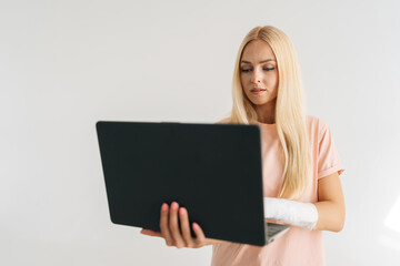 Portrait of thoughtful injured young woman with broken arm wrapped in plaster bandage holding laptop and working typing, standing on white isolated background. Concept of insurance and healthcare.