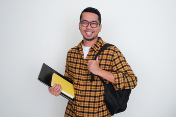 Adult Asian man holding laptop and book with school bag on his back smiling confident