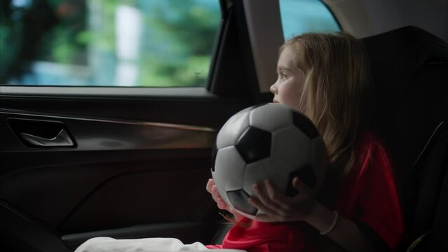 Cute little girl wearing football uniform riding to or from training or match with her parents. Holding a soccer ball in her hands