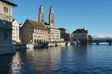 Fototapeta na wymiar View on Limmat River bank surrounded by historical buildings enlighten by sun. In background is Quay Bridge in Zurich in front view and Grossmunster cathedral. 