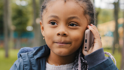 Little ethnic girl talking on mobile phone outdoors portrait funny multiracial child answer telephone call small African American baby kid daughter schoolgirl standing in street talk with smartphone