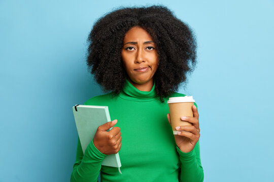 Studio shot of disappointed curly woman stands against blue wall, holds cup of coffee and personal organizer, wears casual green turtleneck