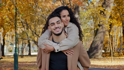 Happy ethnic multiracial Hispanic married couple outdoors cuddle on date in autumn park smiling partners girl hugging guy by shoulders man holding piggyback woman on back romantic date love cuddling